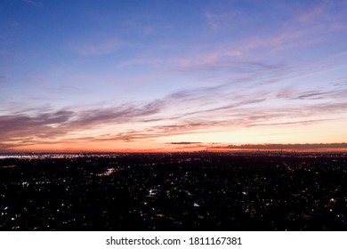 an aerial view of a very colorful sky during sunset. The neighborhood below is dark with some lights on. The drone camera is high enough to see the horizon