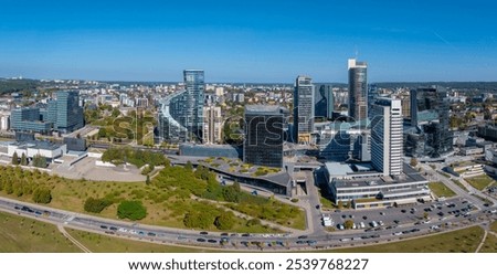 Aerial view of Vilnius, Lithuania, featuring Europa Tower, skyscrapers, and green spaces. A busy road and clear blue sky highlight urban vibrancy.