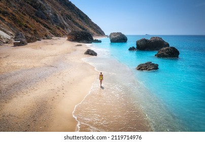 Aerial view of walking young woman on the sandy beach near sea with waves at sunset. Summer vacation in Lefkada island, Greece. Top view of sporty girl, azure water, mountain. Lifestyle and travel