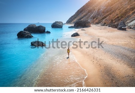 Aerial view of walking young woman on empty sandy beach near sea with waves at sunset. Summer vacation in Lefkada island, Greece. Top view of sporty girl, blue water, mountain. Lifestyle and travel