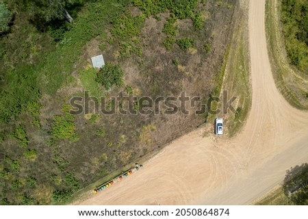 Aerial view of a white car parked on a dirt road beside colorful rubbish bins and the tin roof of an old shed amongst bushland