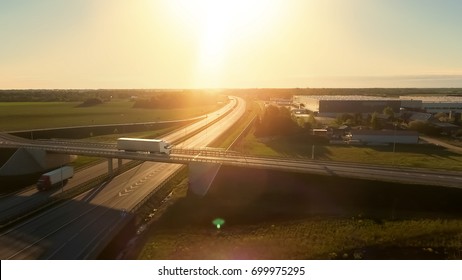 Aerial View Of White Semi Truck With Cargo Trailer Passing Highway Overpass/ Bridge. Eighteen Wheeler Is New, Loading Warehouses Are Seen In The Background.