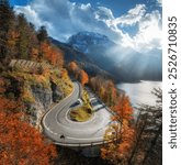 Aerial view of winding mountain road at sunset in autumn. Alps, Italy. Curved alpine road, snowy rocks, trees, red leaves, lake, sky with sunbeams. Colorful. Top view of road on mountain pass in fall