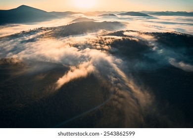 Aerial view of winding road snakes through dense, mist-covered forest in early morning light. Lush green trees with layers of mist creating dreamlike atmosphere as mountains rise in background. - Powered by Shutterstock