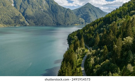 An aerial view of a winding road through dense forest along the edge of a calm fjord in Norway. The surrounding mountains are covered in lush greenery, Fjaerlandsfjorden, Fjord, Vestland, Norway - Powered by Shutterstock