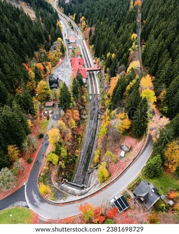 An aerial view of a winding rural road and train tracks, running through a dense forest in autumn