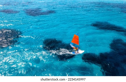 Aerial view of windsurfer on blue sea at sunny summer day. Windsurfing. Extreme sport and vacation. Top view of man on windsurfer board, waves, clear azure water in Sardinia, Italy. Tropical landscape - Powered by Shutterstock