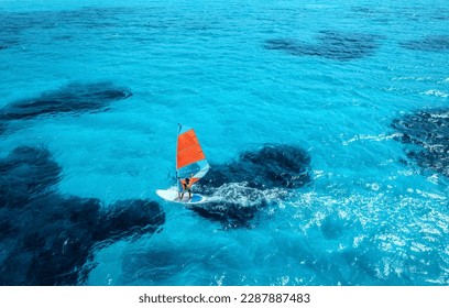 Aerial view of windsurfer on blue sea at sunny summer day. Windsurfing. Extreme sport and vacation. Top view of man on windsurfer board, waves, clear azure water in Sardinia, Italy. Tropical landscape - Powered by Shutterstock