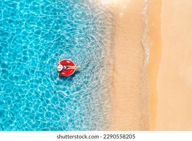 Aerial view of a woman in hat is swimming with red swim ring in blue sea at sunset in summer. Tropical landscape with girl, clear water, waves, sandy beach. Top view. Travel. Sardinia island, Italy	