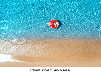Aerial view of a woman in hat swimming with red swim ring in blue sea at sunrise in summer. Tropical landscape with girl, clear water, waves, sandy beach. Top view. Vacation. Sardinia island, Italy	