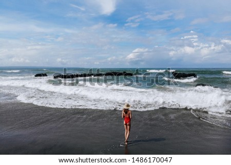 Aerial view of woman in red bikini and straw hat walking on ocean beach with black sand. Vacation in Bali. Photo from drone