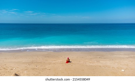 Aerial view of woman in red dress, sitting on the sandy beach, enjoying soft turquoise ocean wave. Tropical sea in summer season on Megali Petra beach on Lefkada island.