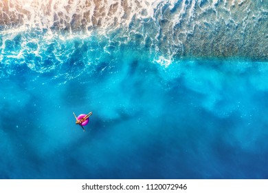 Aerial view of woman swimming on the pink swim ring in the transparent sea and beautiful waves in Europe. Summer landscape with girl, beach, blue water at sunset. Top view. Travel and holiday. Resort