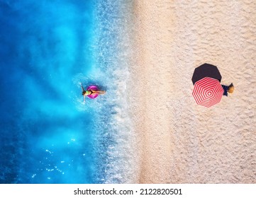 Aerial view of a woman swimming with pink swim ring in blue sea, sandy beach and red umbrella at sunset in summer. Tropical landscape with girl, clear water, waves. Top view. Lefkada island, Greece