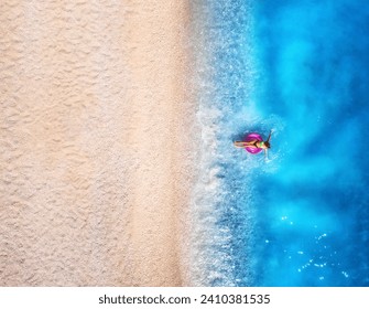 Aerial view of a woman swimming with pink swim ring in blue sea, empty sandy beach at sunset in summer. Tropical landscape with girl, clear water, waves. Top view. Lefkada island, Greece. Background - Powered by Shutterstock