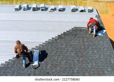 Aerial view in worker hands installing bitumen roof shingles with air hammer and nail - Shutterstock ID 2127758060