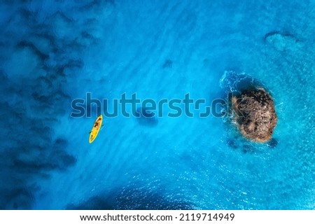Aerial view of yellow kayak in blue sea at sunset in summer. Man on floating canoe in clear azure water, rocks, stones. Lefkada island, Greece. Tropical landscape. Sup board. Active travel. Top view