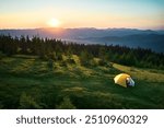 Aerial view of yellow tent on grassy hillside, illuminated by rising sun. Breathtaking panorama of forested mountains and rolling hills under clear sky, creating tranquil and picturesque camping spot.