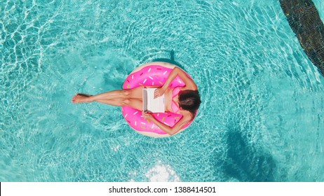 Aerial view of a young brunette woman swimming on an inflatable big donut with a laptop in a transparent turquoise pool - Shutterstock ID 1388414501