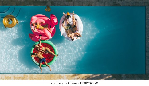 Aerial View Of Young People On Summer Vacation Enjoying On Fun Inflatable Floats In Swimming Pool. Group Of Friends Relaxing On Air Mattresses In Pool.