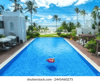 Aerial view of young woman on swimming ring in pool near ocean at sunrise. Top view of girl, blue water, beautiful hotel, palm trees, sandy beach, sea and sky with clouds. Pwani Mchangani, Zanzibar
