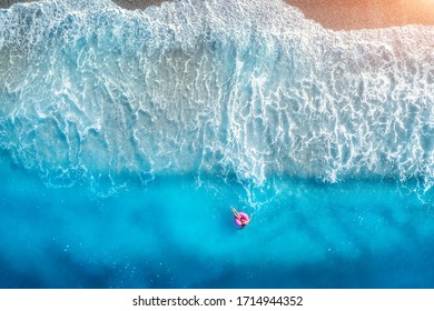 Aerial view of a young woman swimming with the donut swim ring in transparent blue sea with waves at sunset in summer. Tropical landscape with girl, azure water, sandy beach. View from above. Travel