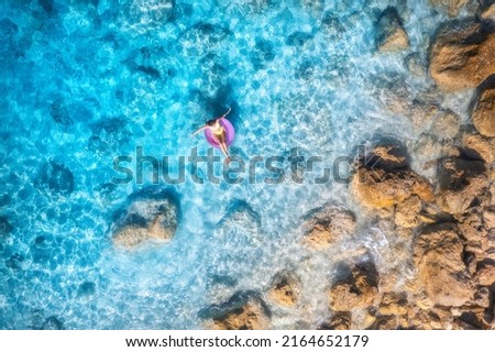 Aerial view of a young woman swimming with pink swim ring in blue sea at sunset in summer. Tropical landscape with girl, clear water, stones, sandy beach. Top view. Vacation in Lefkada island, Greece