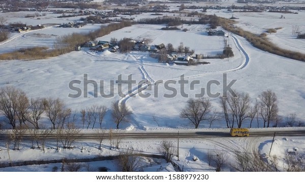 Aerial of the winter road with a\
bus riding along the small village covered by snow. Shot. Winter\
landscape with wooden houses, road and moving yellow\
bus.