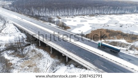 Aerial winter traffic. Drone countryside road. Snowy bridge trough frozen lake asphalted icy danger driveway in cold cloudy day.