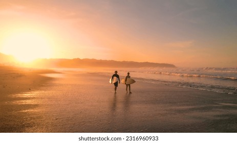 AERIAL: Young couple walking on beautiful sandy beach after sunset surf session. Stunning view of orange glowing sandy beach and rolling ocean waves. Surf couple on a surf trip at exotic Playa Venao.