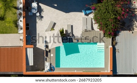 Aerial zenithal view of an empty rectangular pool. The pool is located in the garden of a beautiful house. There are steps to enter the water and loungers surrounding the pool.