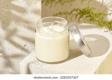 Aesthetic composition with aromatic candle in jar on concrete podium. Mockup soy wax candle in natural style. Scented handmade candle with green leaves.  Handmade spa product  from soy wax in glass