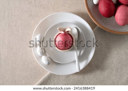 Aesthetic Easter holiday table setting, pink colored egg in cup with bunny ears, on white plates, spoon, small candies, colored eggs on neutral beige linen tablecloth and napkin background.