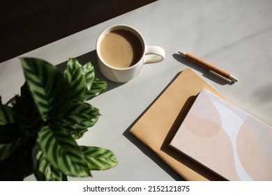 Aesthetic Feminine Workspace. Cup Of Coffee, Notebooks, Green Plant On Home Office Desk Table At Sunset. 