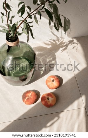 Aesthetic lifestyle still life with peaches, green glass jar with plants and sun light shadows on a beige linen tablecloth background. Summer slow living concept