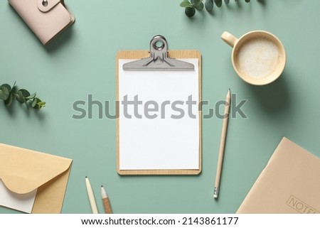 Aesthetic minimal office desk table with clipboard mockup, coffee cup, stationery and eucalyptus leaves on green background. Flat lay, top view.