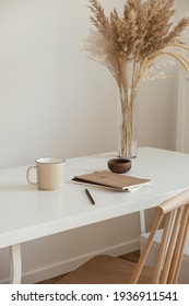 Aesthetic Minimal Office Workspace Interior Design. Mug, Notebook, Pampas Grass Floral Bouquet On White Table Against White Wall. Girl, Woman Boss Work At Home Business Concept.