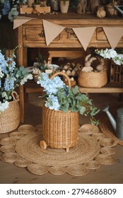 An aesthetic spring composition on a jute rug. A large wicker basket with a lush bouquet of blue delphiniums and roses stands next to a vintage metal watering can.