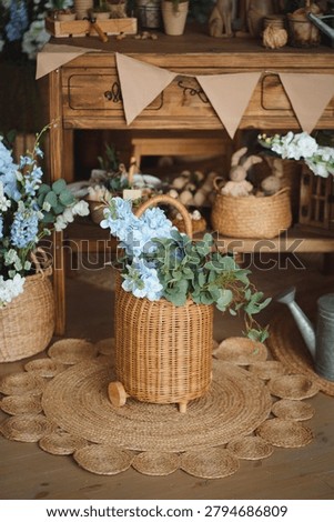 An aesthetic spring composition on a jute rug. A large wicker basket with a lush bouquet of blue delphiniums and roses stands next to a vintage metal watering can.