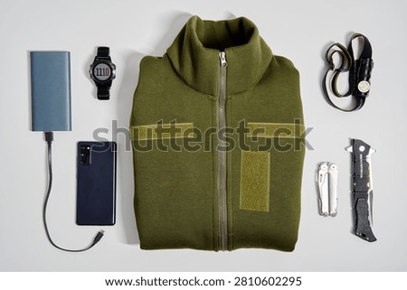 An aesthetically organized flat lay showcasing outdoor gear essentials, including a green jacket, multitools, tech devices, and accessories, perfectly suited for adventure or survival scenarios.