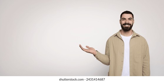 Affable bearded man smiling and gesturing with his hand open as if holding something invisible, dressed in a casual beige shirt and white t-shirt, on a wide light background
