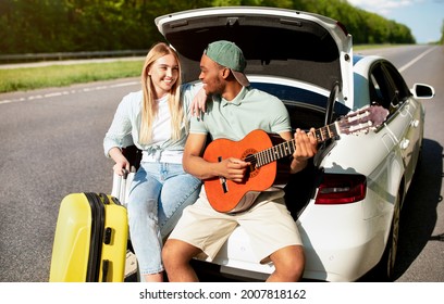 Affecionate Black Guy Playing Guitar For His Girlfriend While Sitting In Car Trunk During Their Travels. Multiracial Millennial Couple Enjoying Road Adventure Together, Having Fun Summer Vacation