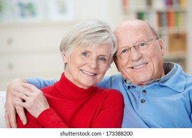 Affectionate attractive elderly couple sitting together on a couch in the living room with their arms around each other smiling at the camera