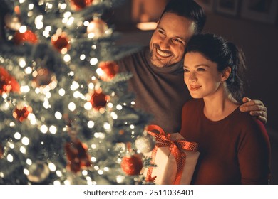 An affectionate beautiful couple enjoying each others company in a loving embrace, celebrating Christmas and admiring the decorated Christmas tree. - Powered by Shutterstock