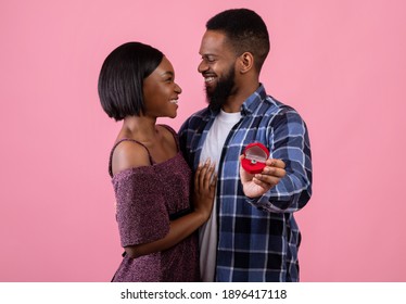 Affectionate Black Couple Hugging And Looking At Each Other, Holding Box With Engagement Ring On Pink Studio Background, Selective Focus. Marriage Proposal, Wedding Concept