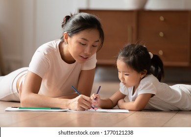 Affectionate Caring Vietnamese Ethnicity Woman Lying On Floor With Cute Small Preschool Biracial Child Daughter, Involved In Hand Drawing Pictures Together In Paper Album, Childcare Hobby Pastime.