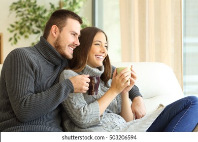 Affectionate Couple Wearing Sweater Holding Hot Coffee Cups Sitting On A Sofa In The Living Room At Home In Winter 