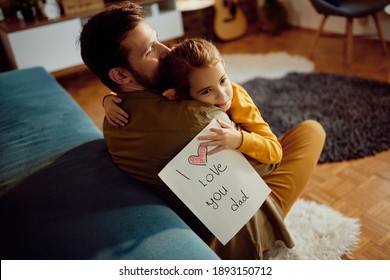 Affectionate daughter embracing her father while giving him drawing and 'I love you dad' inscription at home  