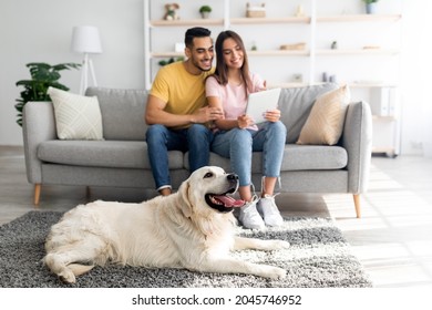 Affectionate Diverse Couple Using Tablet On Couch, Cute Pet Dog Lying On The Floor At Home, Selective Focus. Millennial Family With Touch Pad Relaxing At Sofa In Living Room