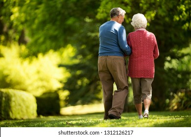 Affectionate Elderly Couple Walk In A Sunny Garden Together.